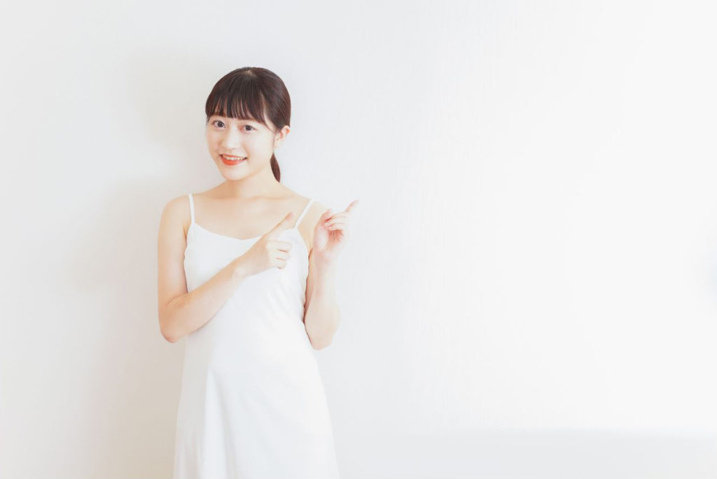 白いワンピースの両手の人差し指を左上に指している笑顔の若い女性
