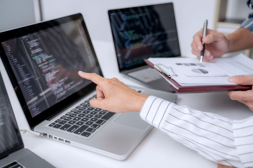 ノートパソコン画面を指さしプログラミングを教えている男性の手と教わってノートを取っている男性のて